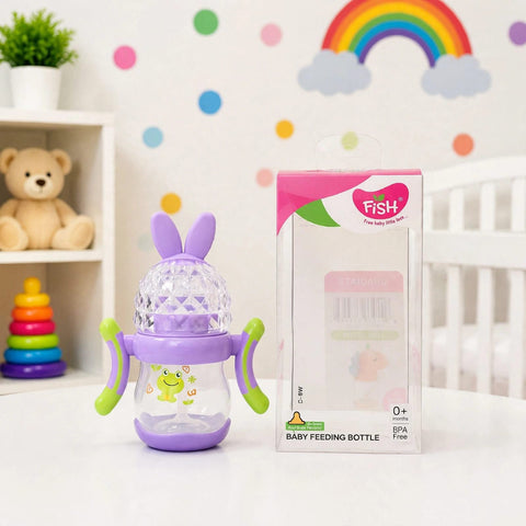 A baby feeding bottle with purple bunny ears and green handles sits next to its retail box on a white table. The bottle has a clear body, purple base and lid, and green side grips.