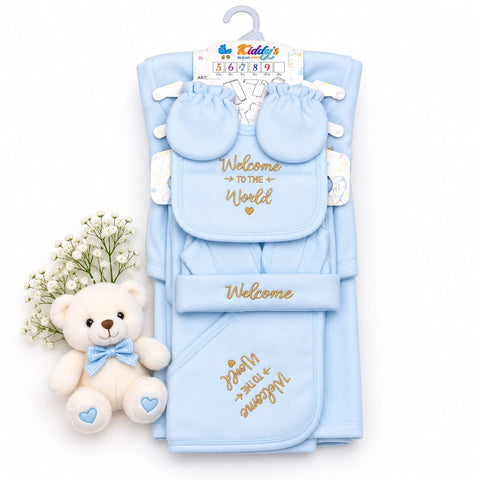 A baby welcome gift set in light blue, including a wrap blanket, bib, booties, and a matching hat, decorated with gold 'Welcome' text. It is displayed on a white background next to a small white teddy bear with a blue bow tie and white baby's breath flowers.
