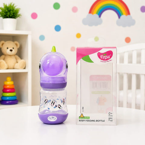 A purple and clear baby feeding bottle with a dinosaur design stands next to its retail box on a white table. The background shows a soft, decorated nursery setting with a teddy bear, rainbow, clouds, and colorful dots.