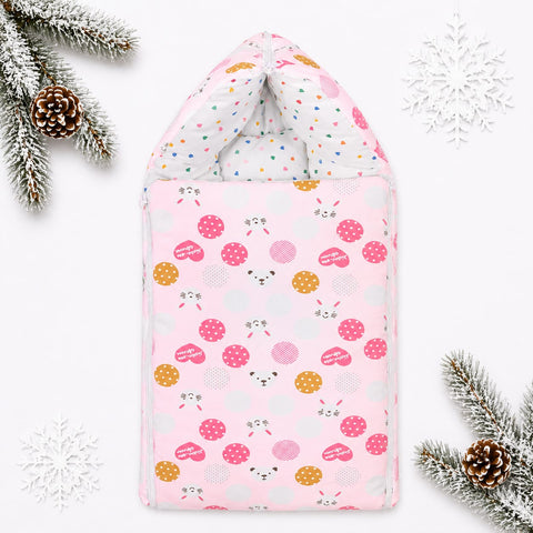 A pink baby blanket or swaddle with a playful pattern of pink and orange hedgehogs, white polka dots, and small animal faces, styled with winter-themed props including pine branches, snowflakes, and a pinecone on a white background.