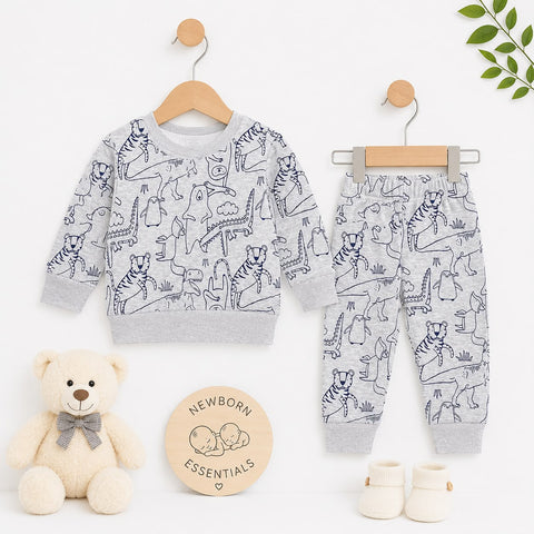 A baby clothing set displayed on wooden hangers against a clean white background, styled with a cream teddy bear, a round 'Newborn Essentials' sign, and small cream booties. The set includes a long-sleeve sweatshirt and matching pants in light heather gray with a dark navy dinosaur and tiger line-art print.