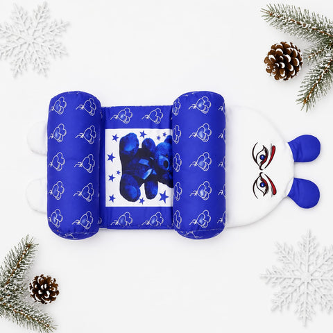 A plush baby support pillow styled in a flat lay with winter-themed props like pine branches, pinecones, and white snowflakes on a white background. The pillow features a blue and white cloud pattern on the sides, a central blue bear graphic, and a white bunny face with blue ears on the right.