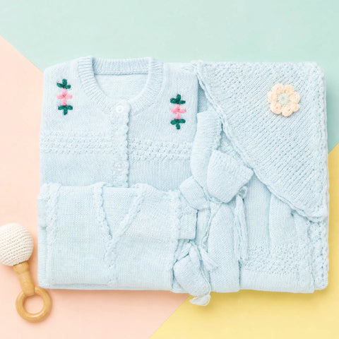 A folded light blue knitted baby outfit set on a pastel geometric background, including a long-sleeve cardigan with embroidered flowers, a matching wrap, and a small wooden teething toy.