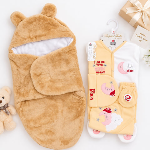 A baby sleep wrap in plush tan faux fur with bear ears and a white lining, displayed next to a matching yellow and white baby bodysuit with cartoon moon graphics and text. The set is styled on a white background with a teddy bear, a gift box, and a small card.
