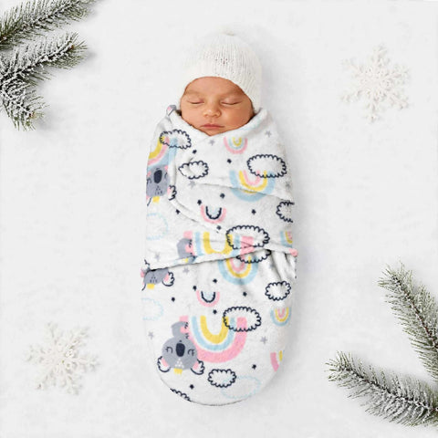 A sleeping newborn baby wrapped in a white swaddle blanket with a colorful cartoon rainbow and cloud pattern, wearing a white knit hat, styled on a white background with snowflake decorations and pine branches.