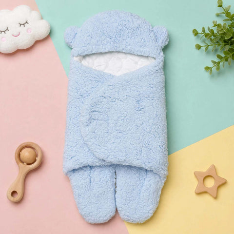 A light blue plush baby swaddle wrap with integrated footies, laid flat on a pastel geometric background with a cloud toy, wooden rattle, and plant leaves around it.