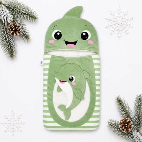 A green plush whale-shaped baby blanket or pouch with a smiling whale face on the hood and a larger whale applique on the front, set against a white background with decorative pine branches and snowflakes.