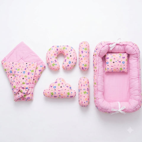 A pink baby bedding set arranged on a white background, including a bassinet, a hooded blanket, a U-shaped pillow, two cylindrical support pillows, and a cloud-shaped pillow. The fabric features a light pink base with a colorful alphabet pattern.