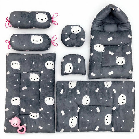 A 6-piece baby bedding set in dark gray with a white cartoon bear and bone pattern, including a changing pad, mattress protector, bassinet pad, pillow, footmuff, and two cylindrical pillow covers with pink ties, plus a pink teething ring.