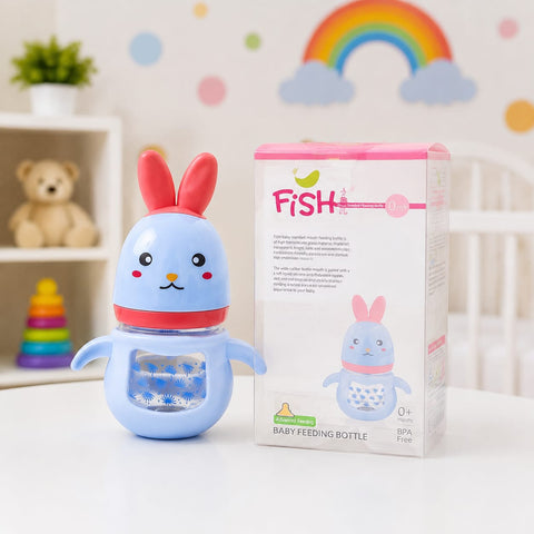 A light blue bunny-shaped baby feeding bottle with red ears and a red collar stands next to its retail box on a white tabletop. The box is labeled 'FISH' and 'Baby Feeding Bottle.