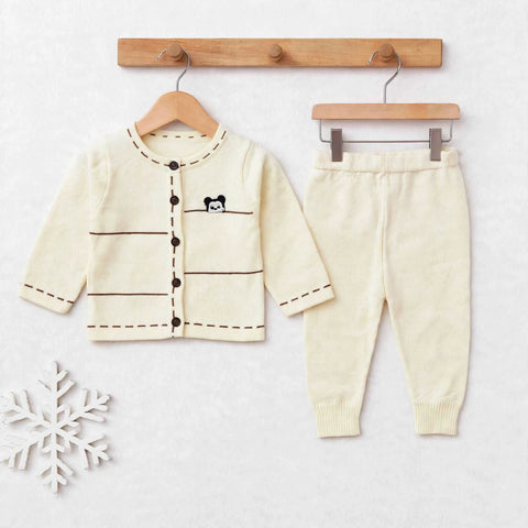 A baby knit two-piece outfit set consisting of a cream-colored cardigan with brown trim and buttons, and matching cream pants, displayed on wooden hangers against a light background with a wooden peg rack and a white snowflake decoration.