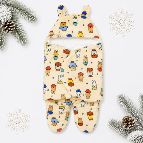 A baby footed swaddle blanket with a hood, featuring a playful multicolor cartoon animal print on a warm cream background. The swaddle is styled in a flat-lay arrangement with winter-themed props including pine branches, pinecones, and white snowflakes on a clean white background.