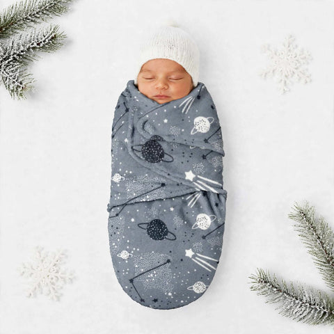 A sleeping newborn baby wrapped in a slate blue swaddle blanket with a space-themed print of planets, stars, and shooting stars, wearing a white knit hat. The scene is styled with white snowflakes and green pine branches on a white background.