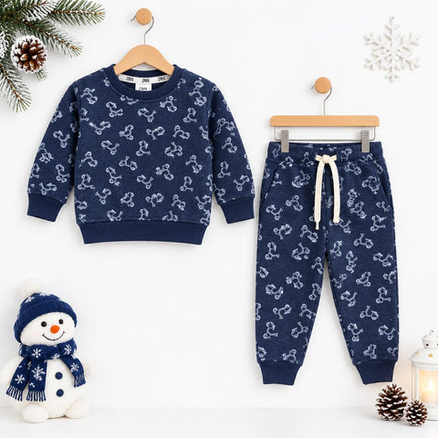 A children's two-piece pajama set hanging on wooden hangers against a white wall, featuring a navy blue sweatshirt and matching joggers with a white bone pattern. The set is styled with winter props including a snowman plush, pinecones, a snowflake ornament, and a small lantern.