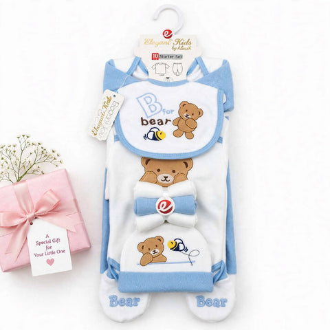A baby footed sleeper set in white and light blue, featuring a teddy bear and bee graphic. The set includes a hooded sleeper, a matching bib, and a small hat, displayed on a hanger next to a pink gift box with a tag.
