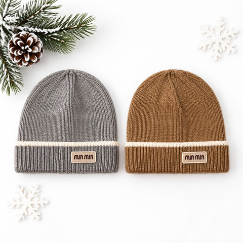 Two ribbed knit beanie hats displayed side by side on a white background with seasonal flat-lay props. The left beanie is heather gray with a cream stripe and a tan 'min min' logo patch.