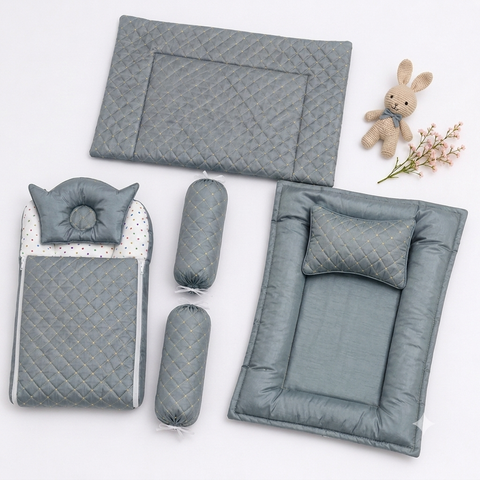 A baby bedding set arranged on a white background, including a quilted mattress protector, a changing pad, a bassinet cover with a pillow, two cylindrical bolster pillows, and a small plush bunny toy with dried flowers.