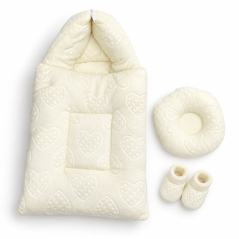 A cream-colored baby bedding set on a white background, including a hooded quilted blanket with an embossed heart pattern, a matching circular cushion, and a pair of soft booties.