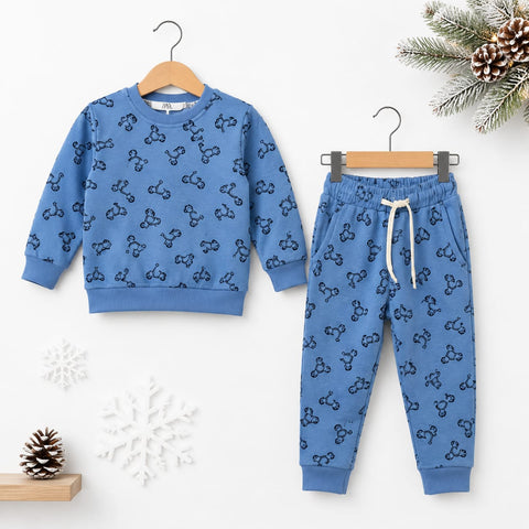 A children's two-piece outfit set featuring a long-sleeve sweatshirt and matching jogger pants in a medium blue color, both printed with a repeating black bow-tie motif. The sweatshirt is shown on a wooden hanger, and the pants are shown on a metal hanger, set against a clean white background decorated with winter-themed props like pine branches, a snowflake, and a pinecone.