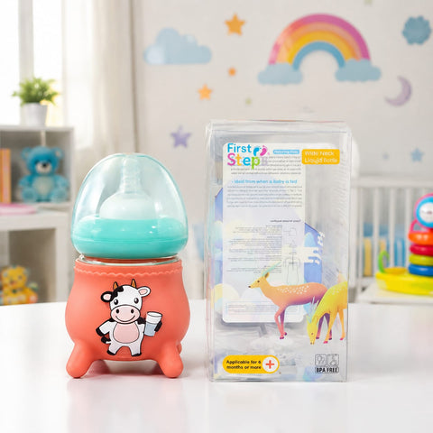 A baby bottle with a translucent teal-blue top and a coral-orange footed base printed with a cow illustration, shown next to its retail box labeled 'First Step' on a white tabletop in a nursery-like setting.