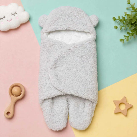 A light gray plush fleece baby footed swaddle wrap with a hood and ear details, laid flat on a pastel-colored background with baby accessories.