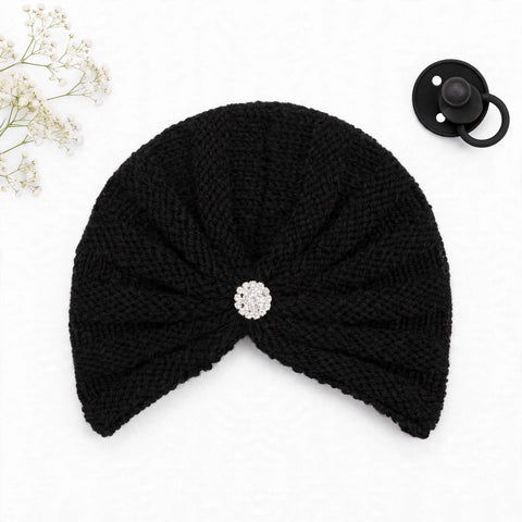 A black knitted baby turban hat with a textured knit pattern and a central silver rhinestone brooch, displayed on a white background with a black pacifier and baby's breath flowers as props.