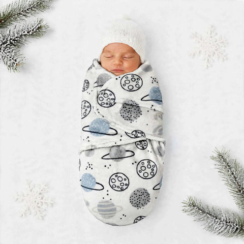 A sleeping newborn baby swaddled in a white wrap blanket with a blue and black space-themed print of planets, moons, and stars, wearing a white knit hat. The baby is photographed from a top-down flat-lay perspective on a white textured background decorated with snowflake graphics and pine branches.