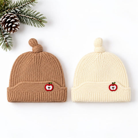 Two ribbed knit baby beanies with pom-poms are shown on a white background, styled with pine branches and a pinecone. One beanie is warm tan/caramel brown, and the other is cream/off-white.