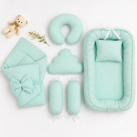 A mint green baby bedding set arranged on a white background, including a bassinet with pillow, quilted blanket with bow, U-shaped cushion, cloud-shaped cushion, two cylindrical bolsters, and a small teddy bear prop.