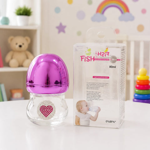 A clear baby bottle with a shiny metallic magenta-purple cap and a rhinestone heart emblem on the front, shown next to its retail box labeled 'Her Fish' on a white tabletop. The background is a softly blurred nursery setting with a teddy bear, shelf, and colorful wall decorations.