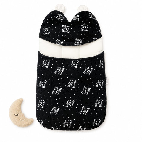 A baby sleep sack with an attached hood in black with a white 'Welcome To World' text and star pattern, featuring a white inner lining and white pom-poms, shown on a white background with a small beige crescent moon toy.