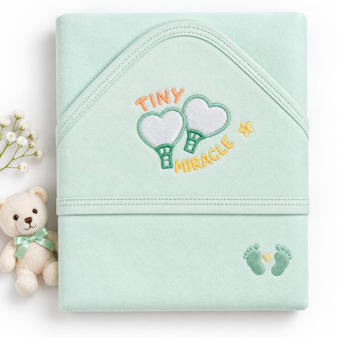 A mint green baby wrap blanket with embroidered 'TINY MIRACLE' text, two hot air balloons, and baby footprints, styled with a small teddy bear and baby's breath on a white background.