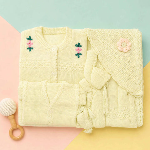 A folded cream-colored knitted baby outfit set, including a cardigan with embroidered flowers, a matching hat, and a small ruffled tie detail, styled on a pastel pink, blue, and yellow background with a wooden rattle prop.