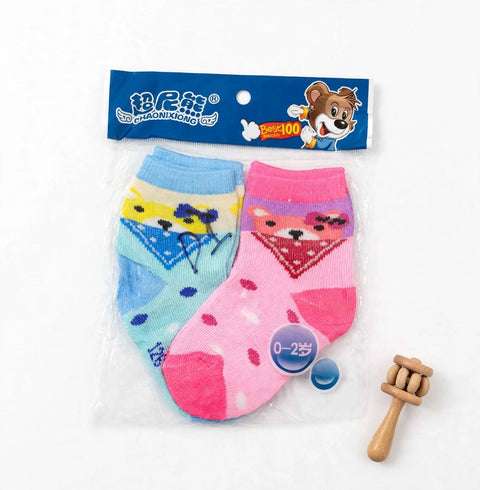 A pair of children's ankle socks in a clear plastic bag with a blue header card. One sock is light blue with a yellow and purple geometric pattern, and the other is pink with a red and purple geometric pattern and small purple dots.