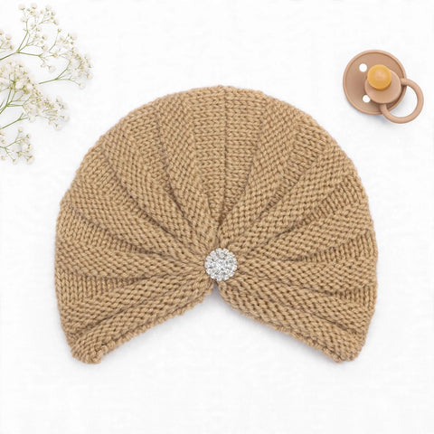 A beige knitted baby turban hat with a textured, ribbed knit pattern and a central silver rhinestone brooch, styled on a white background with baby props including a pacifier and baby's breath flowers.