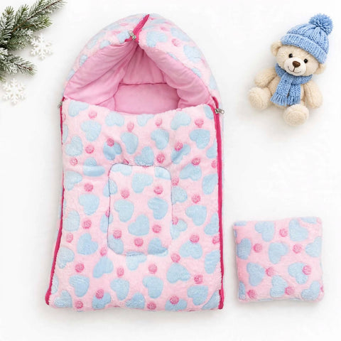 A pink and light blue fleece baby sleeping bag with a heart pattern, shown with its matching zippered pouch on a white background, styled with a small teddy bear and holiday greenery.