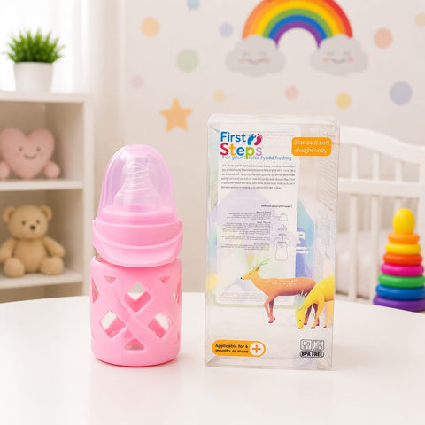 A pink baby bottle with a matching pink protective sleeve stands next to its retail box on a white tabletop. The box is labeled 'First Steps' and shows a deer illustration.