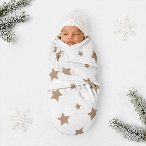 A sleeping newborn baby swaddled in a white fleece blanket with brown star patterns, wearing a matching white knit beanie, laid flat on a white background with decorative snowflakes and pine branches.