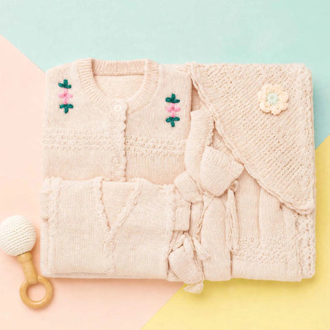 A folded knit baby outfit set in a soft blush pink, including a cardigan with embroidered flowers, a matching wrap or blanket with a flower applique, and a small wooden-handled baby rattle on a pastel geometric background.