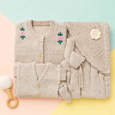 A folded knit baby outfit set in a soft oatmeal beige color, featuring a cardigan with pink and green floral embroidery, a matching knitted bonnet with a flower applique, and a small wooden teething toy, arranged on a pastel geometric background.