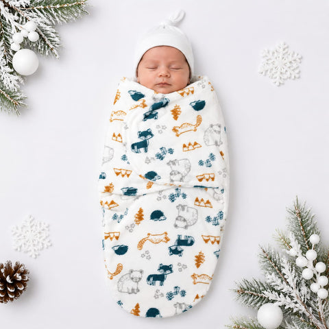 A sleeping newborn baby wrapped in a white swaddle blanket with a colorful forest pattern of moose, foxes, pine trees, and snowflakes, wearing a matching white knit hat. The photo is styled with winter-themed props including pine branches, white ornaments, and snowflakes on a light gray background.