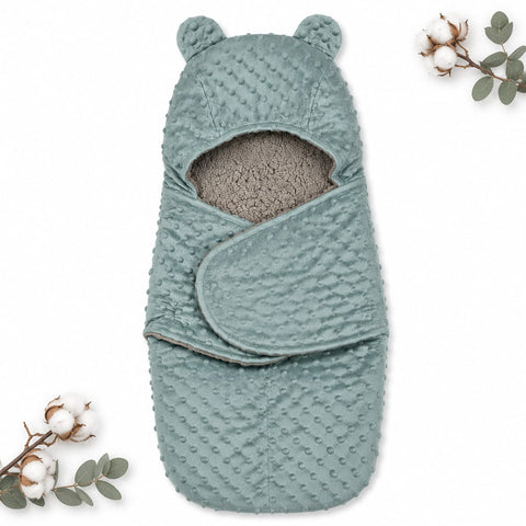 A baby swaddle wrap with a hood featuring bear ears, made of textured dusty blue minky fabric with a soft gray fleece lining. The swaddle is displayed flat on a white background with decorative cotton and eucalyptus sprigs in the corners.