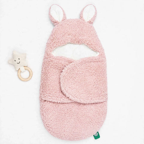 A pink plush baby swaddle blanket with bunny ears and a matching folded cover, shown on a white background next to a small star-shaped wooden teething ring.