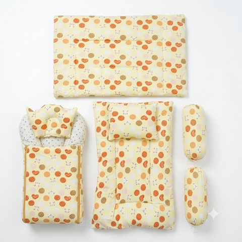 A baby bedding set laid out on a white background, featuring a changing pad, mattress cover, footmuff, and two cylindrical pillow covers, all in a pale yellow fabric with an orange and brown polka dot pattern.