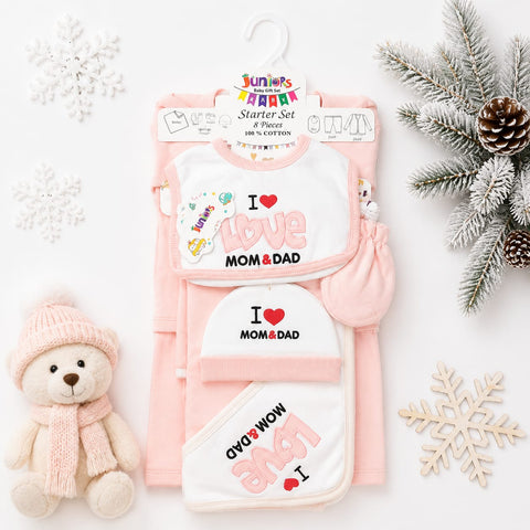 A baby starter set in pink and white, featuring a bib, hat, bootie, and wrap with 'I Love Mom & Dad' graphics, styled with a teddy bear, snowflakes, and pine branches on a white background.