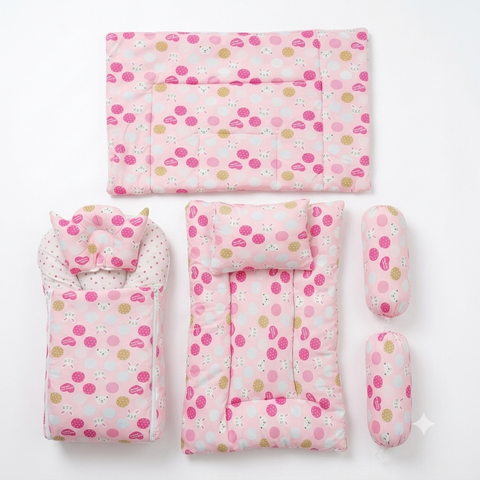 A baby bedding set laid out on a white background, including a quilted mattress protector, a changing pad with a matching pillow, a bassinet cover with a matching pillow, and two cylindrical cushion protectors. The fabric has a light pink base with a pattern of darker pink polka dots, white dots, and small yellow/golden circles.