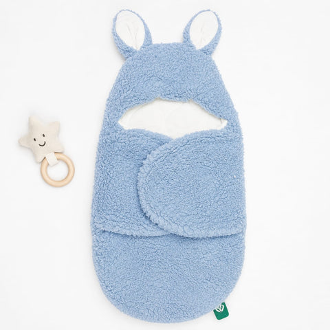 A light blue plush baby swaddle blanket with bunny ears and a matching footed cowl, shown on a white background next to a small star-shaped wooden teether.