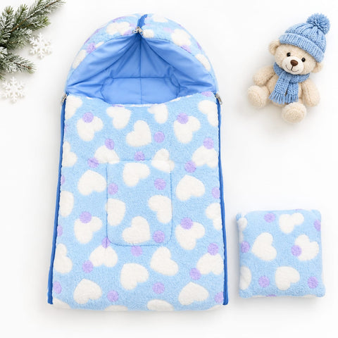 A light blue fleece baby sleeping bag or footmuff with a white and lavender heart pattern, shown open with its hood up and a matching folded pouch beside it. A small teddy bear in a blue knit hat and scarf sits to the right, and evergreen branches with white snowflakes appear in the top left.