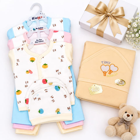 A baby clothing set displayed on a white background, including a cream bib with colorful fruit prints, a light blue top, a pink top, and folded cream and beige garments. A white gift box with a gold ribbon, small white flowers, and a plush teddy bear are arranged around the products.