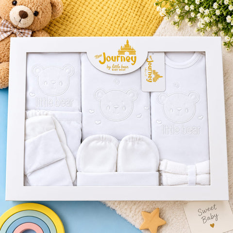 A baby gift set in a white wooden frame, featuring a white long-sleeve bodysuit with an embossed teddy bear face, a matching bib, a pair of footmuffs, and folded white towels. The set is decorated with a yellow 'The Journey by little bear' sticker and a 'Sweet Baby' card, styled with a teddy bear, flowers, and baby accessories.
